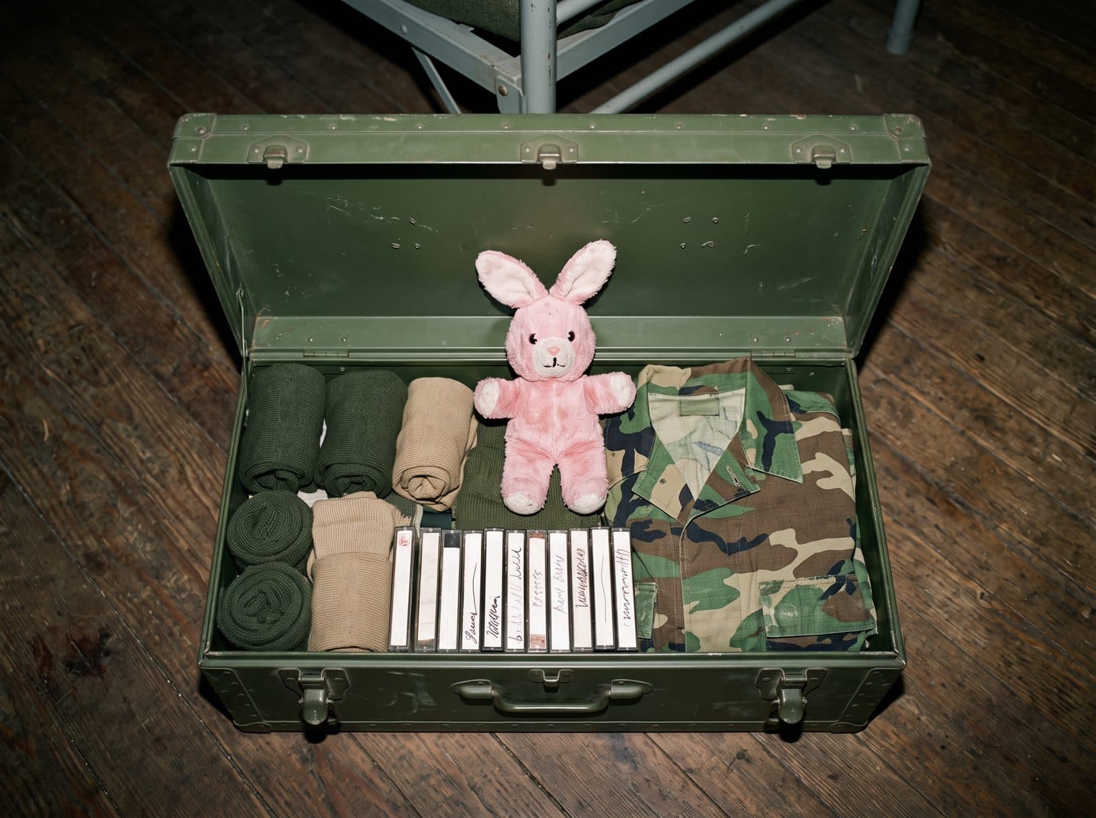 An open army footlocker holding cassette tapes, a stuffed bunny and folded camouflage